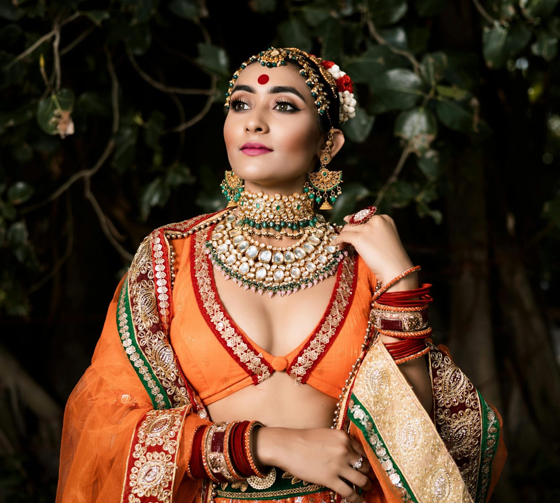 Elegant Indian bride in traditional attire posing outdoors, showcasing exquisite jewelry and vibrant colors.